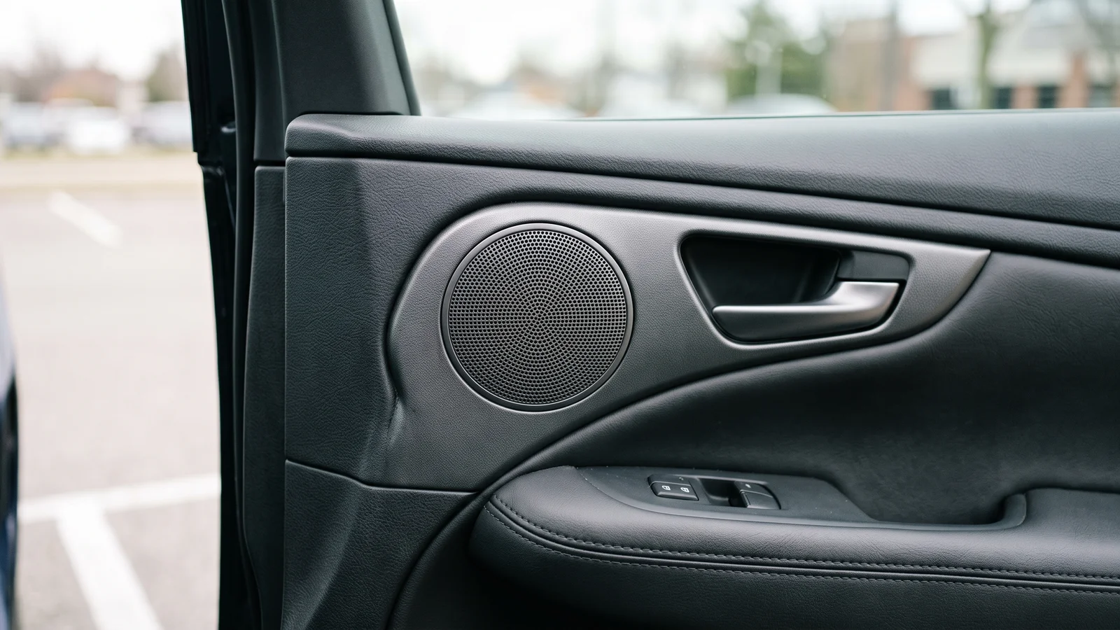 A close-up of a modern car door panel with a factory-installed speaker grille set into the leather-and-plastic trim near the interior handle, illustrating how the Infinity name now lives on mainly as car and OEM audio
