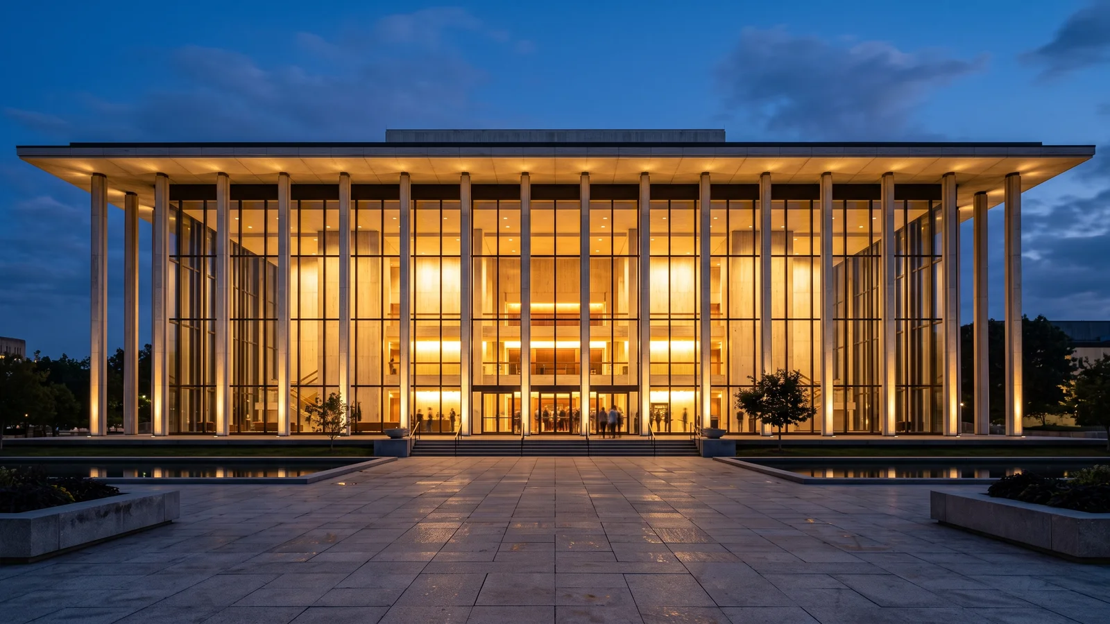 A generic modernist mid-century concert hall exterior at dusk, tall glass facade glowing warm from within, wide plaza in front, evoking a Lincoln Center-style performing arts venue