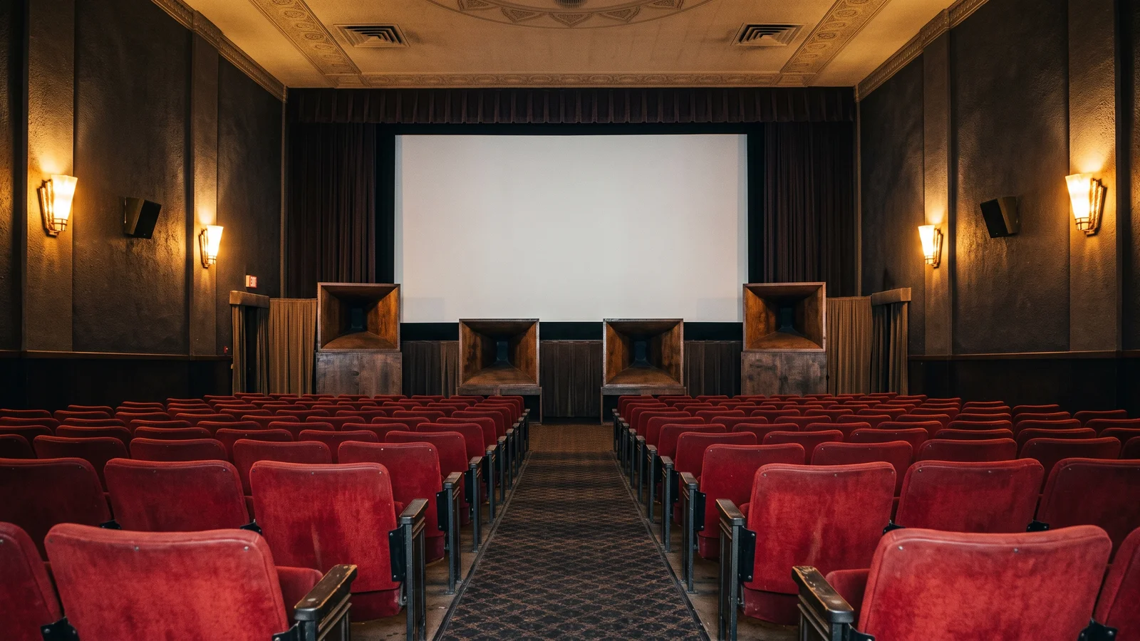 A 1970s movie theater auditorium with large horn subwoofer cabinets standing behind the screen, evoking the Sensurround era